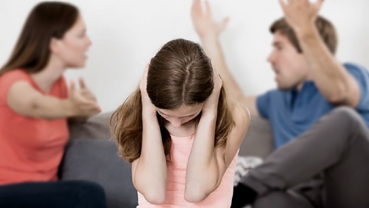 violencia doméstica: imagen en primer plano de una niña tapándose los oídos para no oír la discusión de sus padres detrás de ella