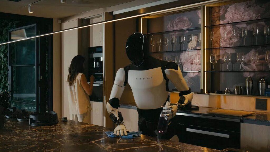 androide optimus de Tesla limpiando la encimera de una cocina mientras una mujer se hace un café al fondo