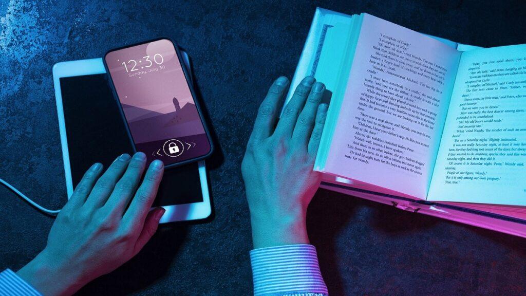 Manos sosteniendo un libro abierto mientras interactúan con un smartphone y una tableta, explorando diversas apps de lectura accesible. La pantalla del smartphone muestra la hora y la fecha con un bloqueo activo. Iluminación en tonos azul y rosa que crea un contraste visual atractivo.