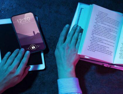 Manos sosteniendo un libro abierto mientras interactúan con un smartphone y una tableta, explorando diversas apps de lectura accesible. La pantalla del smartphone muestra la hora y la fecha con un bloqueo activo. Iluminación en tonos azul y rosa que crea un contraste visual atractivo.