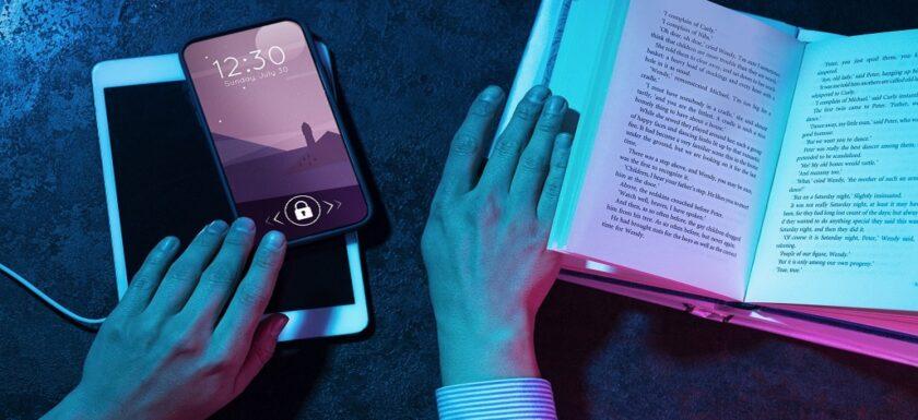 Manos sosteniendo un libro abierto mientras interactúan con un smartphone y una tableta, explorando diversas apps de lectura accesible. La pantalla del smartphone muestra la hora y la fecha con un bloqueo activo. Iluminación en tonos azul y rosa que crea un contraste visual atractivo.