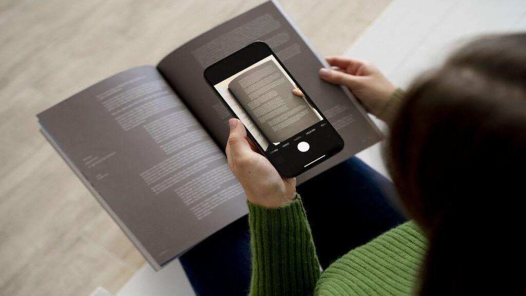 Persona sosteniendo un smartphone sobre un libro abierto, utilizando una app de lectura accesible para digitalizar y mejorar la accesibilidad del texto impreso. La pantalla del smartphone muestra la interfaz de cámara, capturando una página del libro. La escena ilustra la integración de la tecnología con la lectura tradicional.