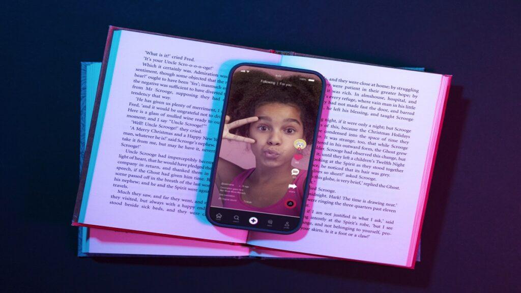 Smartphone colocado sobre un libro abierto, mostrando una app de lectura accesible en la pantalla. El texto del libro parece pertenecer a 'A Christmas Carol' de Charles Dickens, con nombres como Scrooge y Fred visibles. La escena destaca la combinación de lectura tradicional con tecnología accesible.